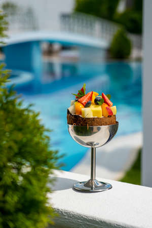 An exotic fruit cocktail in a chrome high-stemmed glass at a luxury resort with a blurred pool in the background. Foreshortening through vegetationの写真素材