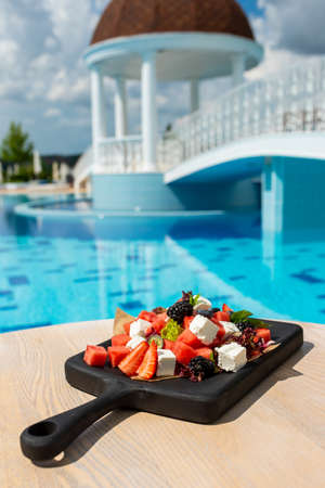 Salad with fruits, vegetables, cheese on a black wooden board. Luxury resort with pool viewの写真素材