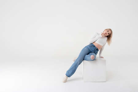 Young beautiful blonde in a white sweater and jeans on a white isolated background sits sideways. Positive and confident personの写真素材