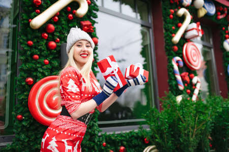 Beautiful girl wearing Santa Claus hat holding Christmas gift boxes in a festive outdoor atmosphere....