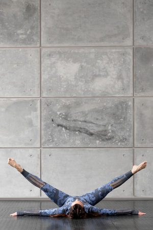 Beautiful sports fitness girl with pigtails. Doing yoga. Lies with legs raised in a twine, against the background of a gray stone wall. Camouflage blue tracksuit. Free space for text. Sport and healthの写真素材