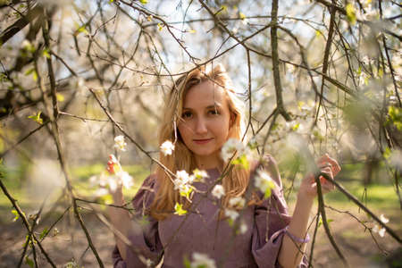 Beautiful girl in a dress looks into the camera through the branches of a flowering tree. spring...