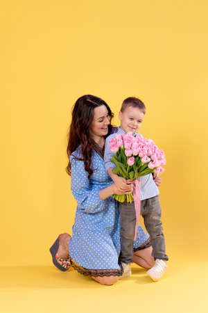 Beautiful mother with a little boy on a yellow background. Loving caring mother plays with her preschool son. A boy with a big bouquet of flowers. child developmentの写真素材