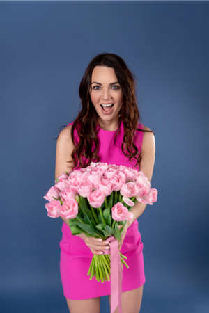 Beautiful brunette with flowers. A woman wears a stylish pink dress, poses on a blue background, expresses happiness. spring conceptの写真素材