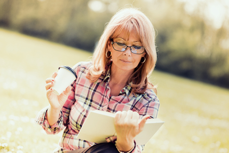 Happy mature woman reading a book during springtime in nature.の写真素材