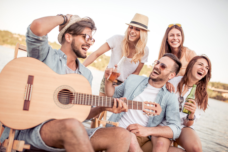 Group of young people listening to friend,playing guitar outdoors.Young friends drinking beers and enjoying music.の写真素材