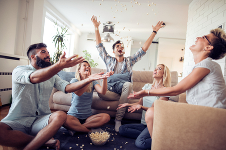 Five happy friends joking and throwing popcorn sitting on a sofa in the living room at home.の写真素材