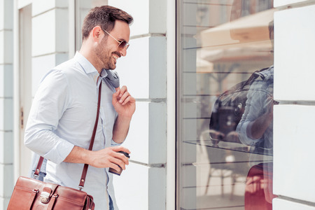 Handsome young man standing on the street, holding coffee to go and watching the showroom.の写真素材