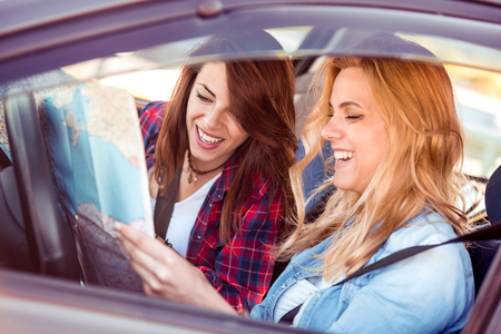 Adventure begins right now! Side view of two beautiful young cheerful women looking away with smile while sitting in car.の写真素材
