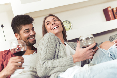 Happy couple sitting on sofa, watching comedy movie and drinking wine.の写真素材