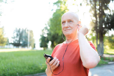 Smiling old man in casual wear and white headphones listening music from his smartphone in city park...