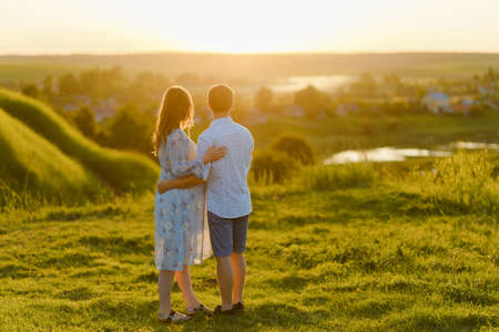 A young beautiful pregnant woman in a long dress and her husband standing together in the hill at...