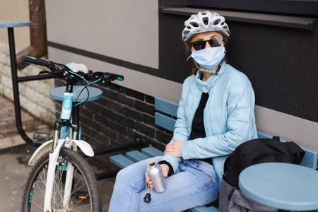A girl cyclist in a helmet and sunglasses and medical mask is sitting at a table and going to drink water.の写真素材