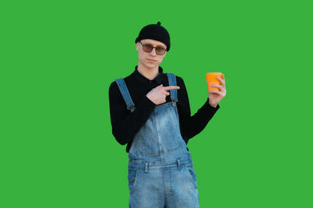 Happy young man in casual clothes on a background of green wall shows thumb up and holds eco cup and...