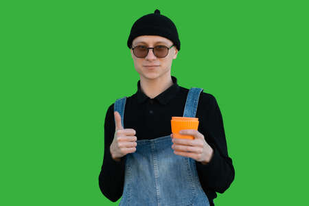 Happy young man in casual clothes on a background of green wall shows thumb up and holds eco cup and smiles while looking at the cameraの写真素材