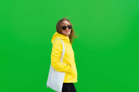 Smiling redhead woman in a raincoat and sunglasses is carrying a reusable pouch isolated on a green...
