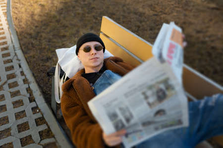 A lying man on a bench is reading a newspaper outdoors.の写真素材