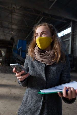 A young woman in a yellow mask checks the technical condition of the enterprise. The woman is using a smartphone and holding sheets of paperの写真素材