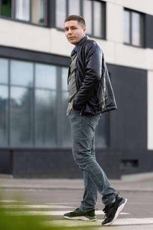 Portrait of a man in a black jacket and jeans looking at the camera crossing the street. street...