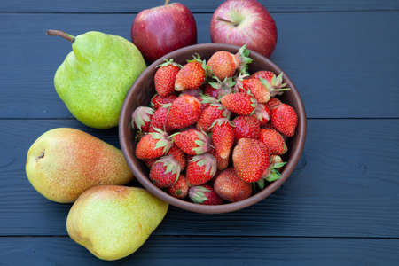 Pears, red apples and ripe strawberries in a clay cup on a dark wooden background. Still life. View...