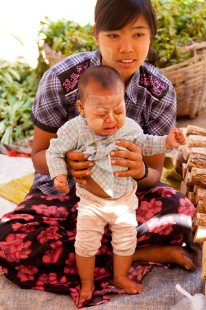 Bagan, Myanmar - February 26, 2011 : Young woman and her baby boy sitting on the ground selling goods in a local market. On their faces they have applied Thanaka - a traditional make up made by mixing ground thanaka root and water. It is used both for sunのeditorial素材