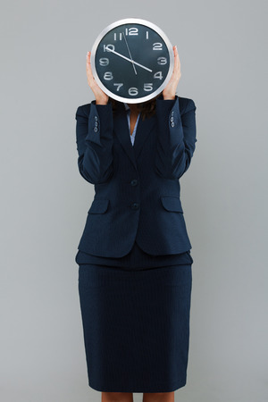 Businesswoman holdng a clock in front of her face isolated on gray backgroundの写真素材