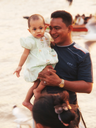 Yangon, Myanmar - February 24, 2011 : Burmese young man holding his daughter in hands on the pier of Yangon, enjoying the hungry seagulls at sunsetのeditorial素材
