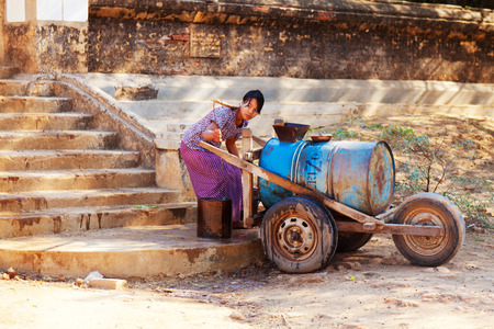 Bagan, Myanmar - February 26, 2011 : Old burmese woman filling up a water tank with buckets of water collected from the pond behind.のeditorial素材
