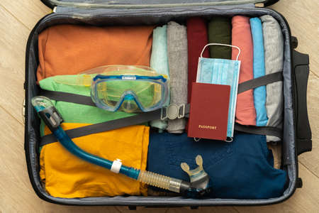 Top view of a suitcase prepared for a seaside holiday during a pandemic. Clothes neatly folded,...