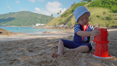 The kid independently plays with a bucket in the sand during a family vacation. A child plays in the sand on the shore of a tropical island during the vacations. family summer holidaysの写真素材