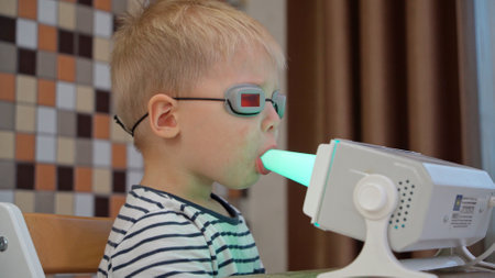 A sick blond boy doing inhalation manipulations treats a cold of the throat at home. Parents teach their children to take care of their health from a young ageの写真素材