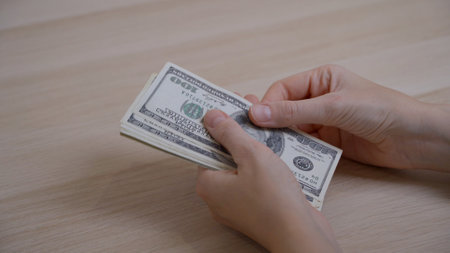 Women's hands hold hundred dollar bills and methodically count them, neatly stacking them in one...