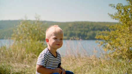 A little blond boy sits in the grass, against the backdrop of natureの写真素材