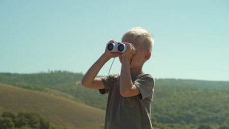 Against the backdrop of nature and the blue sky is a boy. He looks through the binoculars, which is in his hands. The boy is watching what is happening somewhere far away from himの写真素材