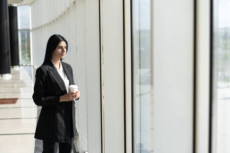 young woman standing near window, looking at city, resting from the work, thinking of something. wide angleの写真素材