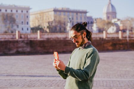 Handsome bearded male hipster making photo for blog sharing positive emotions feeling excited by sunny morning while walking to work using modern smartphone and 4G internetの写真素材