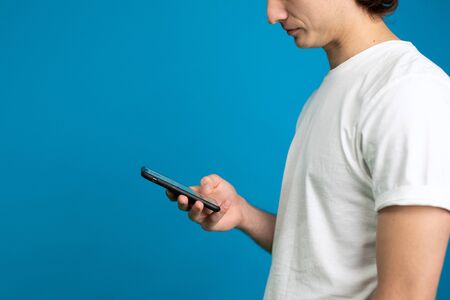 Partial view of young man in white t-shirt using smartphone isolated on blue background.