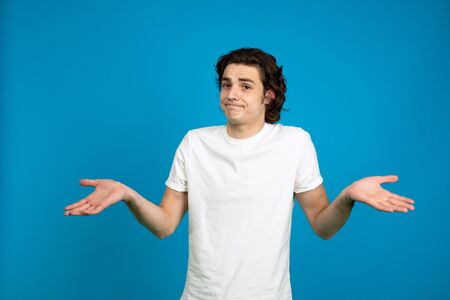 Front view of young man in white t-shirt shrugging isolated on blue background.