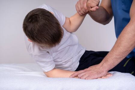The boy sits on a special table, next to him is a doctor in blue uniform and he helps the guy to do physical exercises, rehabilitation conceptの写真素材