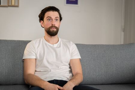 a man sits on a sofa at home, alone, and watches news.  Amazed man reacts to pandemic situationの写真素材
