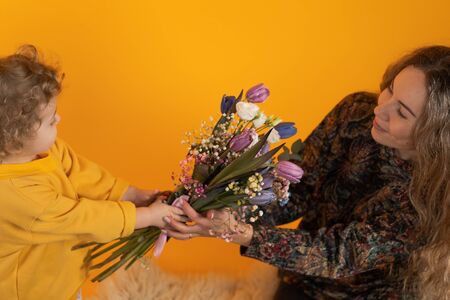 Cute little boy with curly hair holds in his hands a bouquet with multi-colored tulips and gives them to his mother, yellow background, the concept of a holiday and giftsの写真素材