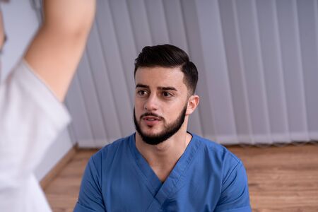 Young pretty doctor in blue uniform and a beard stares at his patient, the concept of health and...