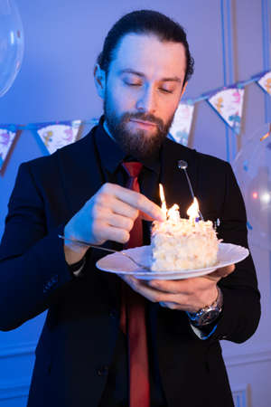 A man holds a piece of cake with candles and looks at it. Dressed in a suit with a tie.