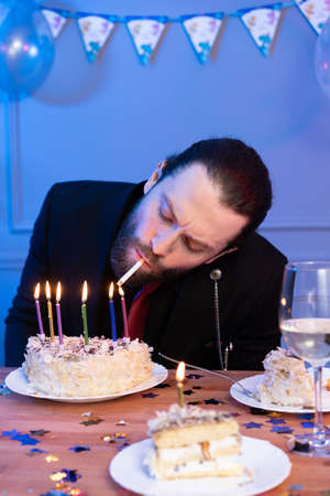 A man lights a cigarette with candles on a cake. Looking at the cake and the cigarette.