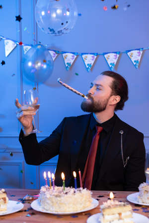 A man sits at a table holding a pipe in his mouth and looks at a glass. Birthday cake on the table.