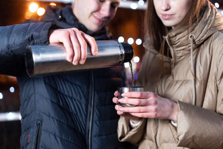 The guy pours tea to the girl in a cup. The girl holds a cup in her hands. Hands in winter.