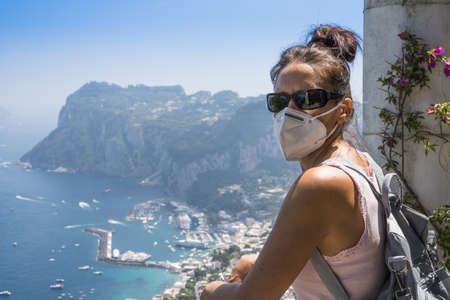 Pretty woman with mask and sunglasses, looking at camera with a beautiful seascape on a sunny summer...