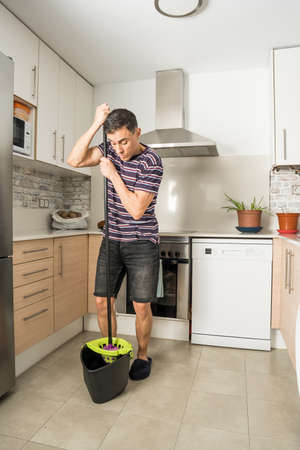 Man in the kitchen wearing casual clothes and slippers, wringing the mop into the bucket. Full body.