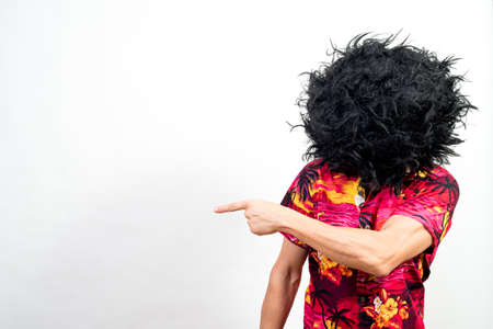 Man with afro wig covering his entire face and Hawaiian shirt, pointing aside. Mid shot. White...