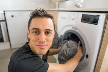 Smiling man putting clothes in the washing machine, in the kitchen. Close up.の写真素材